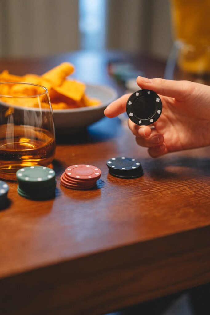 Dubibet Official Entry – Fast Login and 24/7 Access. Close-up of a hand holding a poker chip with whiskey and snacks on the table.